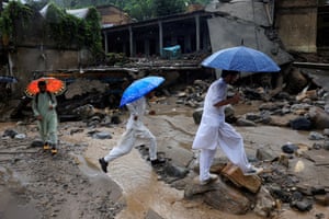 Three men with umbrellas walk beside flood-damaged houses