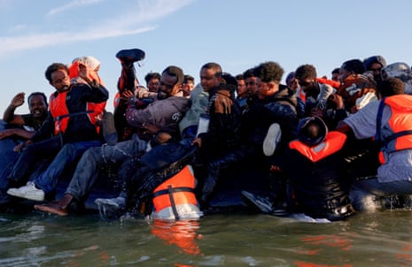 People sit crammed on an overloaded inflatable dinghy; some wear orange life vests