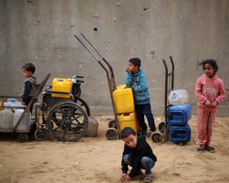 Children with yellow water canisters on trolleys and a wheelchair at a refugee camp