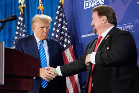 Two men in black suits shake hands in front of American flags.