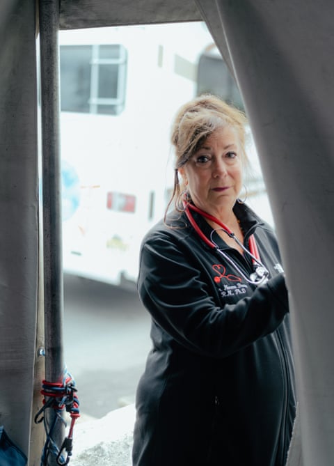 A woman in all black, with a stethoscope around her neck, pulls back the flap of a pop-up tent to look at the camera.