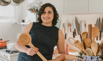 Samin Nosrat, with curly shoulder-length hair and in a sleeveless dress, grins as she leans on a kitchen counter holding a long wooden spoon