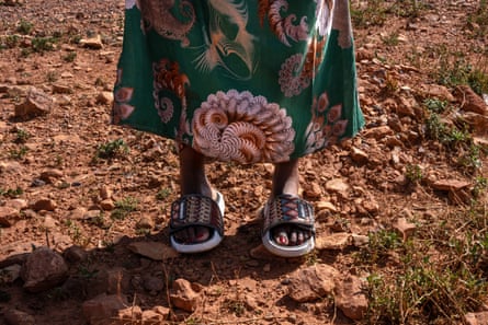 A woman’s feet in sandals stand on red, rocky ground.