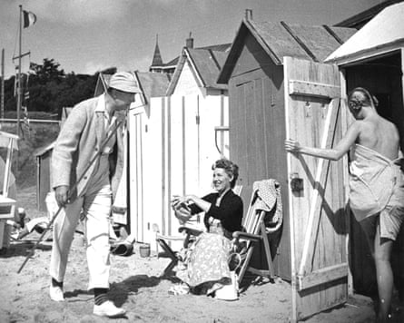 A man in a white suit with a cane and lightweight hat walks among beach huts, with a woman draped in a towel entering one and a woman in a skirt seated in front of another