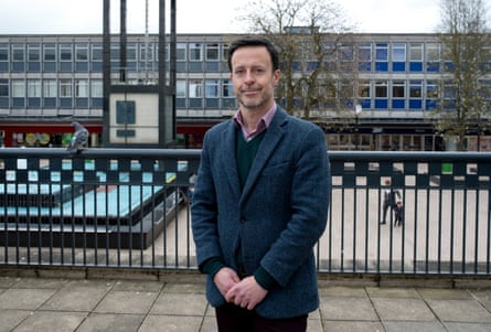 Kevin Bonavia stands on a stone terrace in Stevenage town centre, with 1950s or 60s-style buildings behind him. He is in his late 40s with short dark hair and grey stubble, and wears a dark grey tweed jacket.