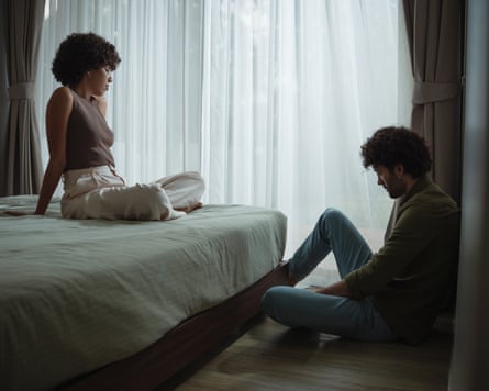 A woman sitting cross-legged on a bed, looking down at a man sitting on the floor with his head down
