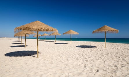 Straw umbrellas on empty white sand beach with clear sea behind, Ilha do Farol, Culatra barrier island, Olhao