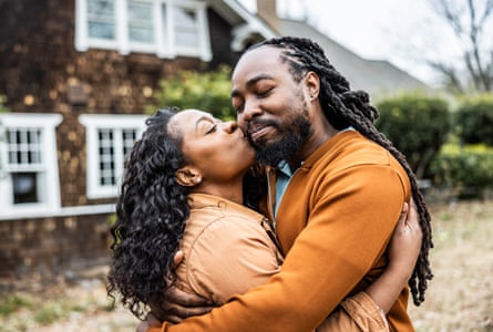Attractive Black couple in front of a nice 1930s house, she is kissing his cheek, he smiles