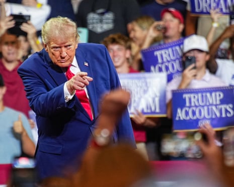 a man in blue suit points to a crowd as people hold signs that say 'Trump accounts'