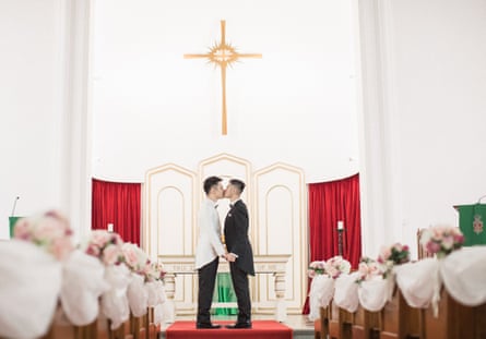 they are photographed in profile in what appears to be a church with white-painted walls and a gold cross and altar in the background; there are pews to either side of the aisle with bunches of flowers at the end.