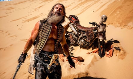 Chris Hemsworth as Dementus brandishing a gun in a desert, in front of a bike modified with the dummy bust of a woman on the handlebars