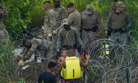 A group of men in olive uniforms stand on one side of loops upon loops of razor wife, while a young man wearing a yellow life jacket, seen from behind, holding a toddler not wearing a shirt, who looks over his shoulder, touches the razor wire with his hand.