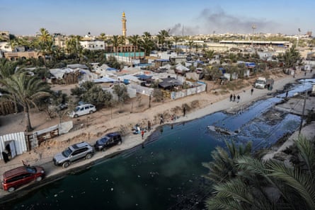 Temporary shelters for displaced Palestinians along a street covered with stagnant wastewater. Smoke rises in the distance