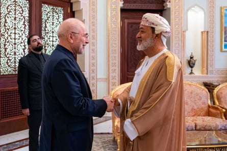 Mohammad Bagher Ghalibaf, wearing a black suit, shakes hands with Oman's sultan, who wears a gold-trimmed, light brown robe-type garment and embroidered headdress. They are standing in an ornately decorated room.
