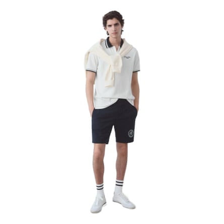 A model wears a white polo shirt with navy detail, a cream jumper draped around the neck, navy knee-length shorts, white socks with navy detail and white tennis shoes.