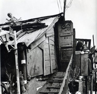 A doll is visible on the roof of a rickety looking home outside which a woman holds her child