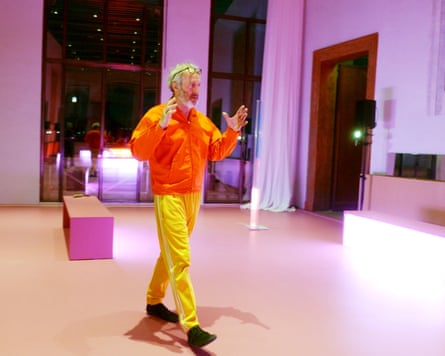 A man in a zip-up sports jacket and tracksuit bottoms walks across a floor, his hands raised in gesture, with an escalator behind him