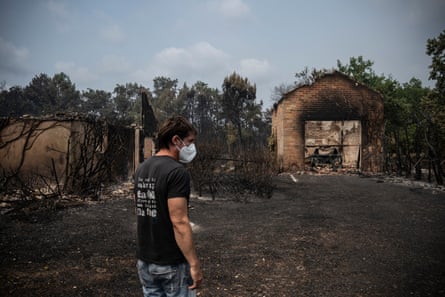 A man wearing a face mask surveys a charred landscape and two burnt-out buildings