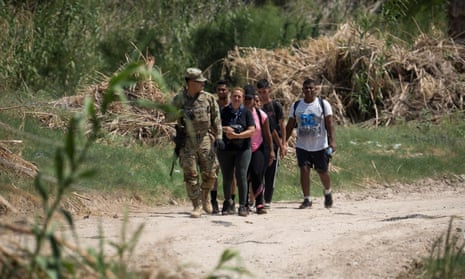 A soldier in army fatigues leads a small group of people wearing backpacks along a dirt road.