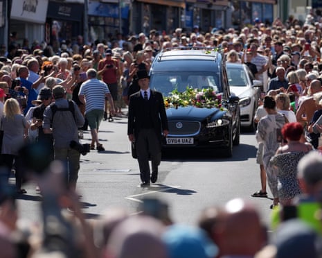 A hearse covered in flowers is driven through a crowd of people