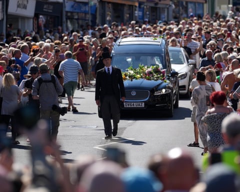 A hearse covered in flowers is driven through a crowd of people