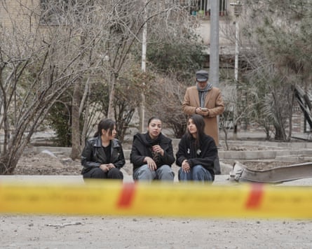 Three teenage girls sit by the roadside as a man stands behind checking his phone. Yellow tape, blurred in the foreground, covers the bottom part of the picture