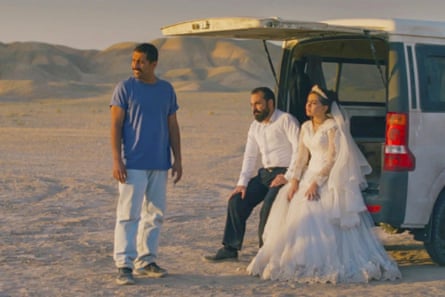 A man standing in a desert next to a woman and man in bridal outfits sitting at the back of a van.