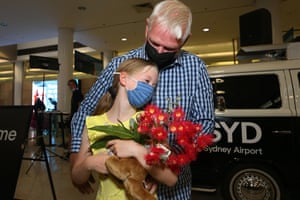 A young girl with blonde hair is wearing a yellow shirt and holding a bouquet of red flowers, her grandfather is wearing a blue check shirt and is hugging her from behind. Charlotte Roempke greets her grandfather Bernie Edmonds as he arrives at Sydney’s international airport