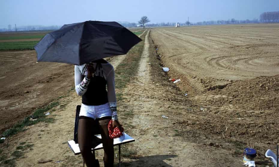 A woman waits in the shade of an umbrella by the edge of a big field for passing clients