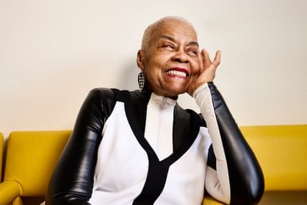 A woman with cropped white/grey hair pictured from the waist up, wearing a black and white high-collared zip-up top and white leaf-shaped earrings, sitting on a mustard-coloured soft, leaning her head on her left hand, with her arm bent at the elbow and leaning on an arm rest, smiling off camera