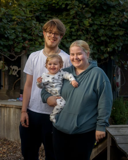 A smiling young woman in a hoodie holding a baby in a babygrow next to a young man