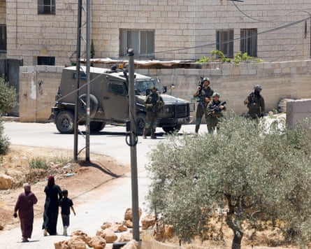 Two Palestinian women and a child walk up a road towards a junction guarded by armed troops and a vehicle