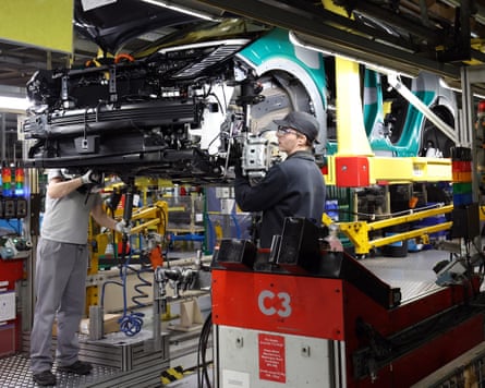 Factory employees, one in foreground with protective glasses and a black baseball cap, working on a raised car in factory.