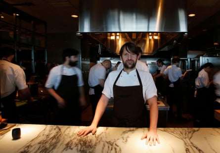 A chef in a kitchen with a dark blue apron with his hands on a wooden table. A group of chefs are behind him