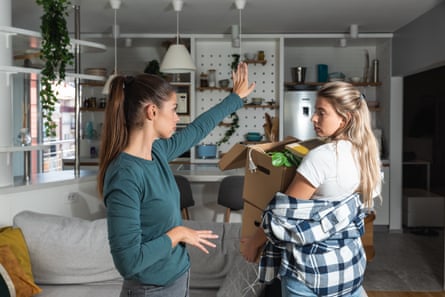 A young woman holding boxes full of stuff in an apartment as she leaves while a woman next to her gestures angrily