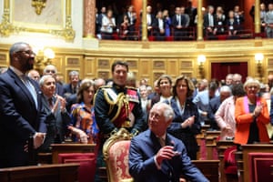 Charles looks to his right as he sits in a chair, as people stand in the senate behind him