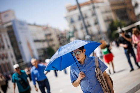 A man in glasses holds a blue umbrella while walking in a sunny Madrid square