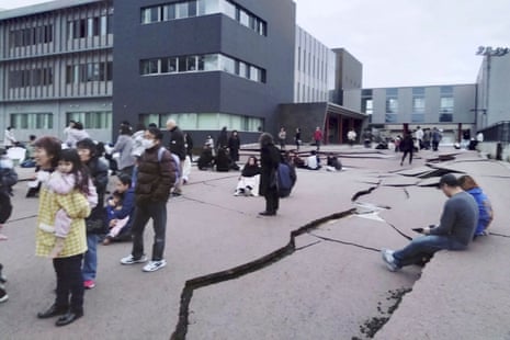 People stand near cracks in tarsealed ground next to multi-storey buildings