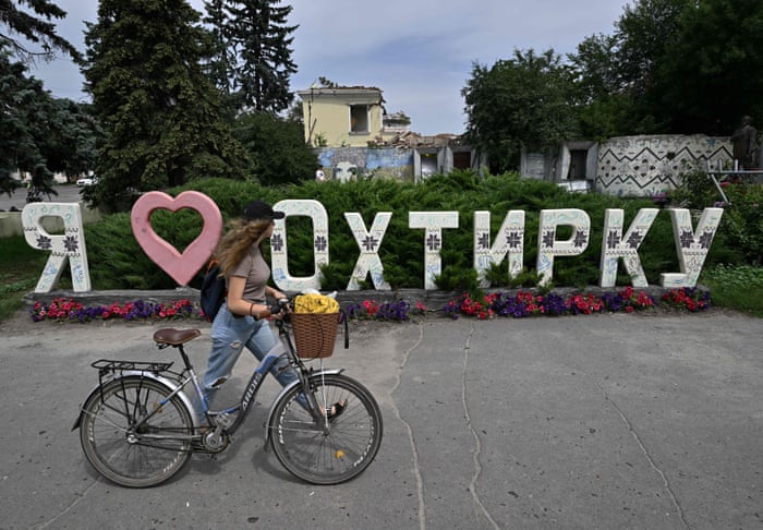 A woman pushes her bicycle past a sign which reads I love Okhtyrka in front of the destroyed city hall in the city.