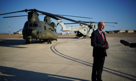 John Healey speaking to journalists, out of shot, on the tarmac of an airbase with helicopters in the background