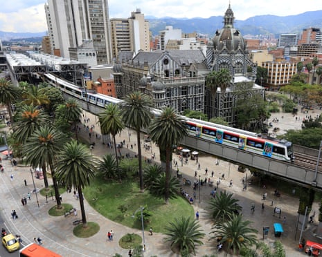 Aerial view of plaza with elevated train running through it, with buildings, trees and lawns