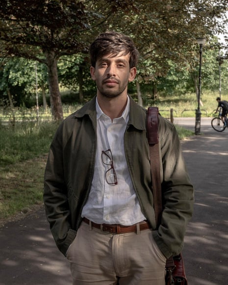 A young man with a beard in a white shirt and jacket with spectacles and a park behind him