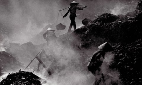 Black and white image of people walking and working on piles of rubbish, while smoke rises around them