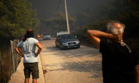 Two men stand with their faces covered watching cars drive away from a fire