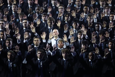 Rows of young men in dark suits and ties raise one hand high in unison