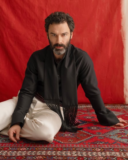 Aidan Turner kneels on a patterned rug leaning on his left hand with his left arm out to his side, wearing a black shirt with fringed black scarf and cream trousers, against a red backdrop