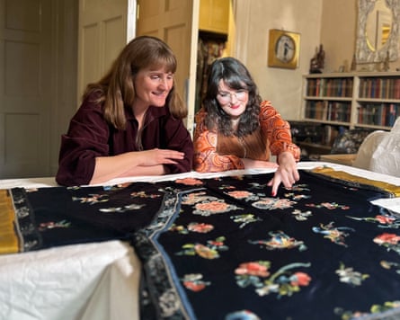 Two women lean over a white-clothed table to examine an intricate, dark-colored textile laid out before them. The fabric is decorated with vibrant, embroidered floral patterns in shades of red, pink, and orange