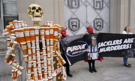 A sculpture of a human figure made of old opioid prescription pill bottles and topped by a fake skull is seen beside protesters dressed as devils. The costumed protesters are holding a black banner that reads: "Purdue American Cartel" and "Sacklers Are Murderers!" The banner also bears an image of man in a tie and white lab coat looking out from behind prison bars.