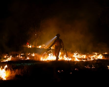 A member of the emergency military wearing a head-torch and carrying a can of fuel walks between lines of flames. He is near-silhouetted in the dark with the flames glowing against the black backdrop.