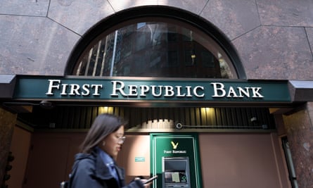 Seorang wanita melihat teleponnya berjalan melewati fasad hijau dan emas dari cabang First Republic Bank